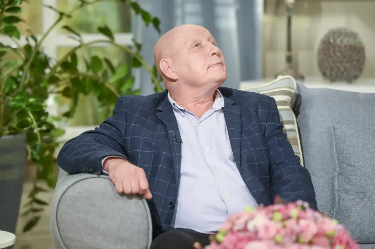Krzysztof Jackowski w okrytym kratkę garniturze, siedzi na kanapie, jakby zastanawiał się nad najnowszymi przepowiedniami.