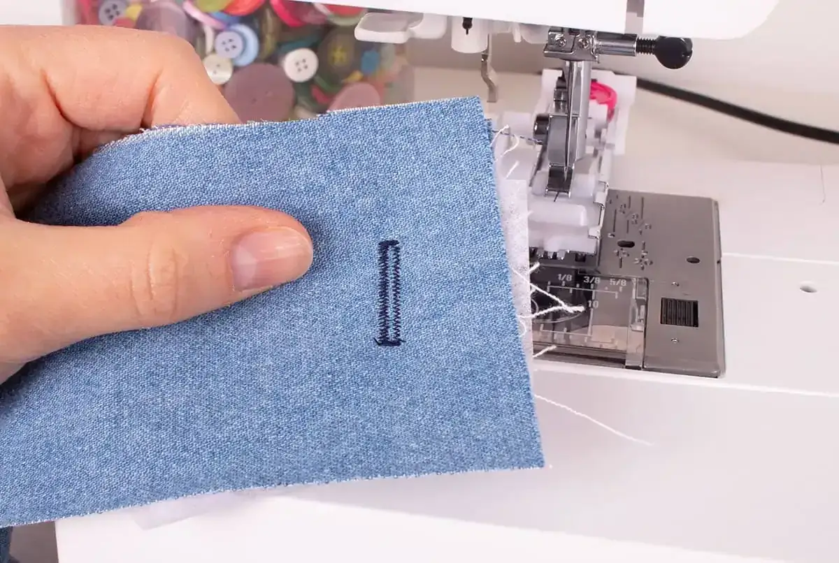 A hand holds denim fabric with a newly sewn buttonhole, demonstrating how to sew a buttonhole on a sewing machine.