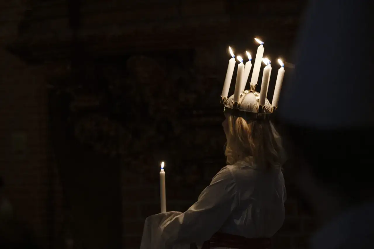 En flicka med ljuskrona bär ett ljus. Hon är klädd i vitt och har ett rött band runt midjan. En vacker bild av storkyrkan lucia.
