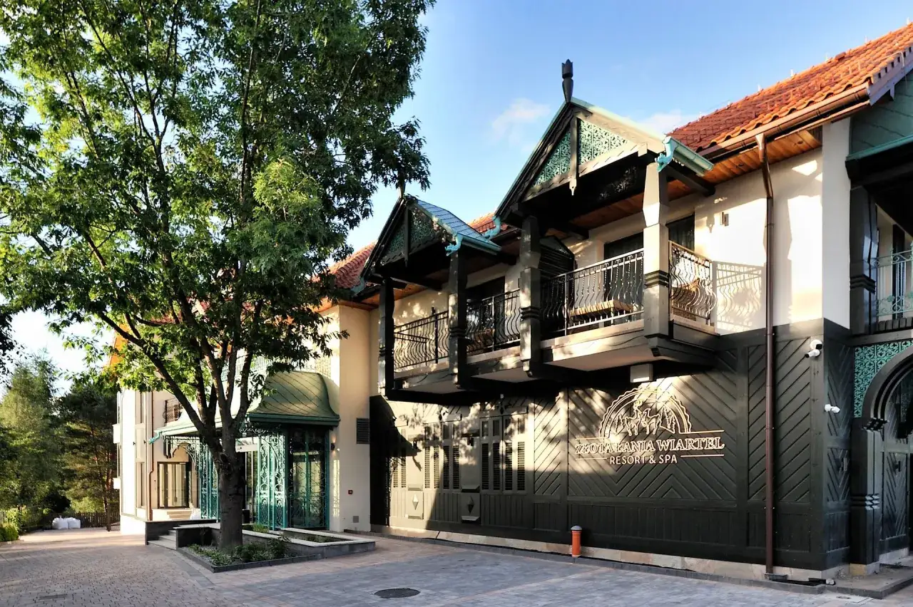 Elegancki budynek Złota Łania Wiartel Resort & Spa z ozdobnymi balkonami i zielonymi elementami architektonicznymi, otoczony drzewami.