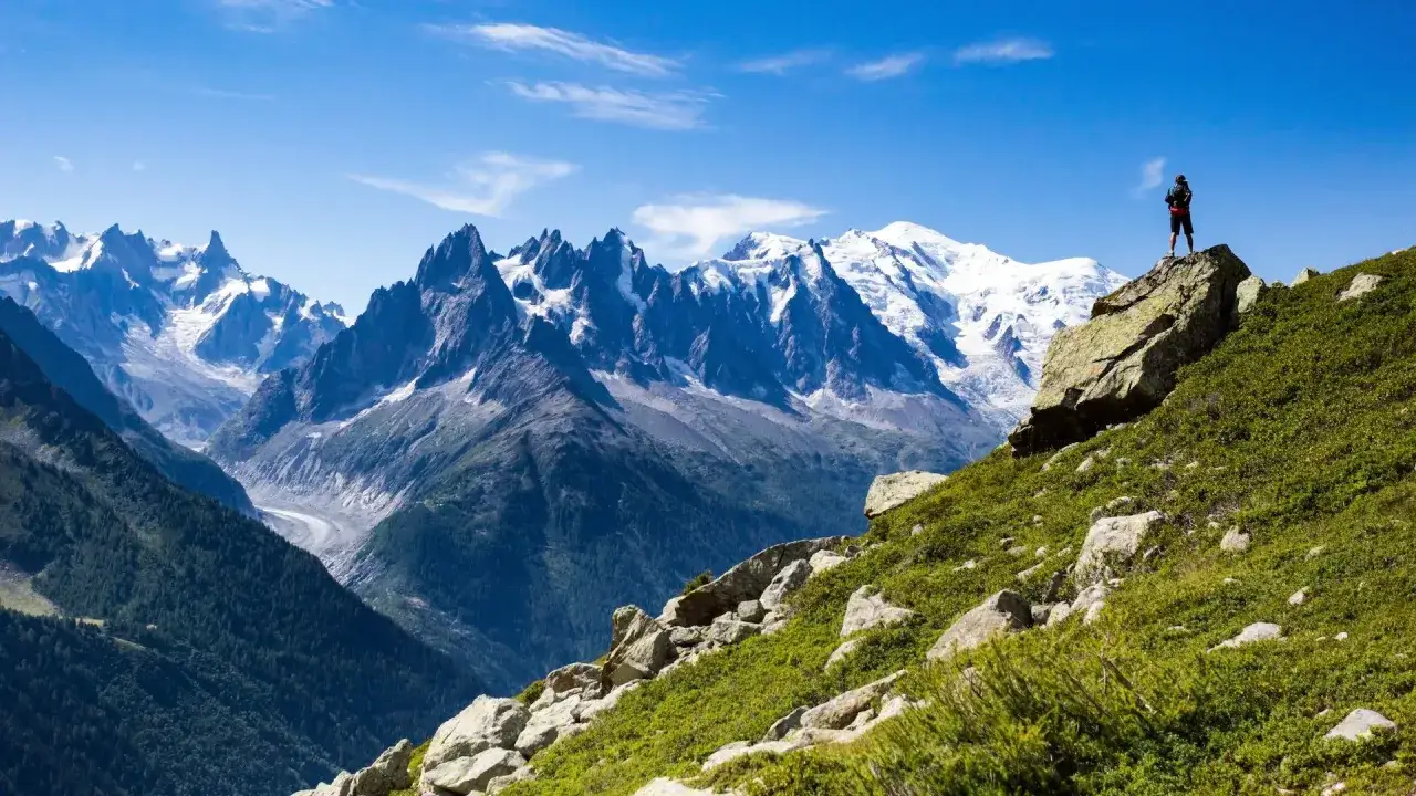 A lone hiker stands atop a rocky outcrop, surveying the majestic Mont Blanc massif during a breathtaking hike.