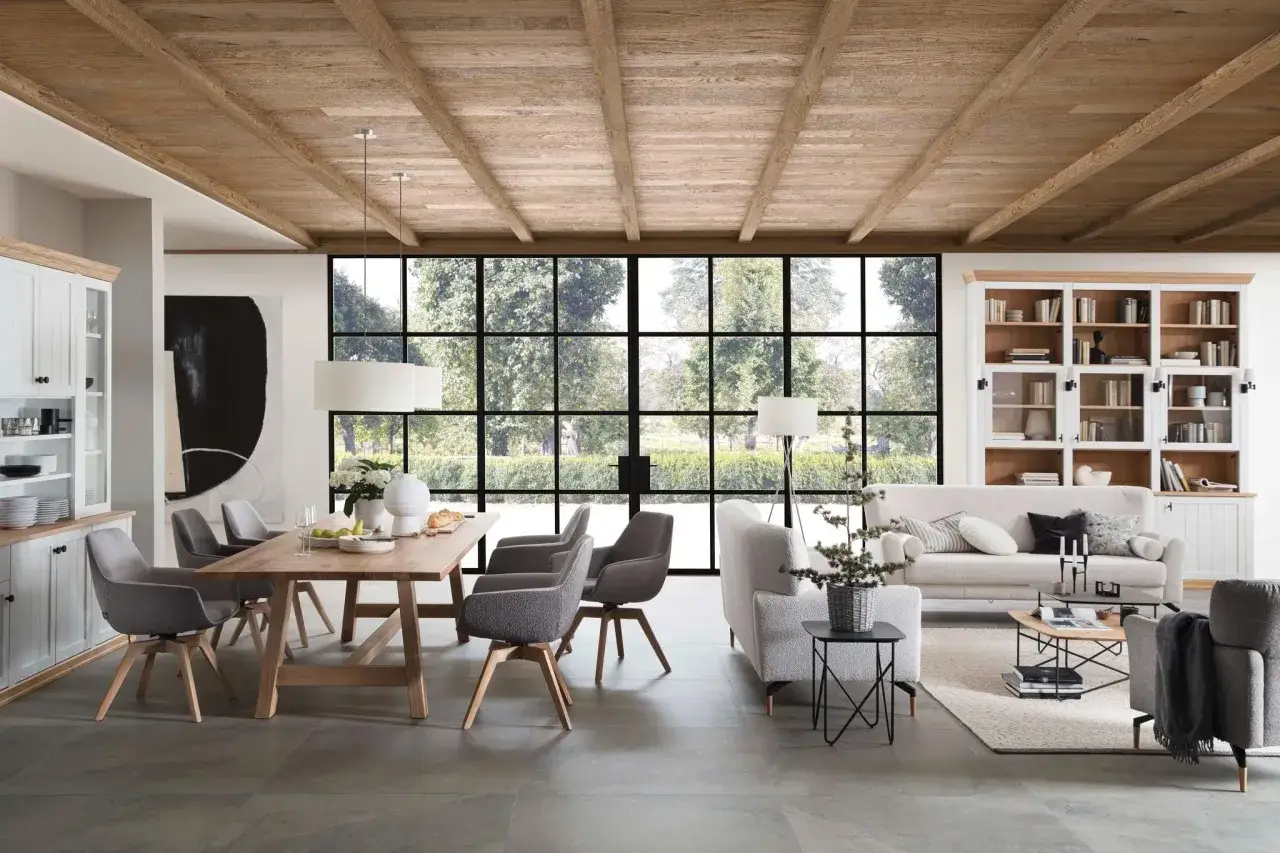 Ein heller Wohn-Essbereich im modernen Landhausstil mit Holzbalkendecke, großem Esstisch, gemütlicher Couch und Bücherregal.