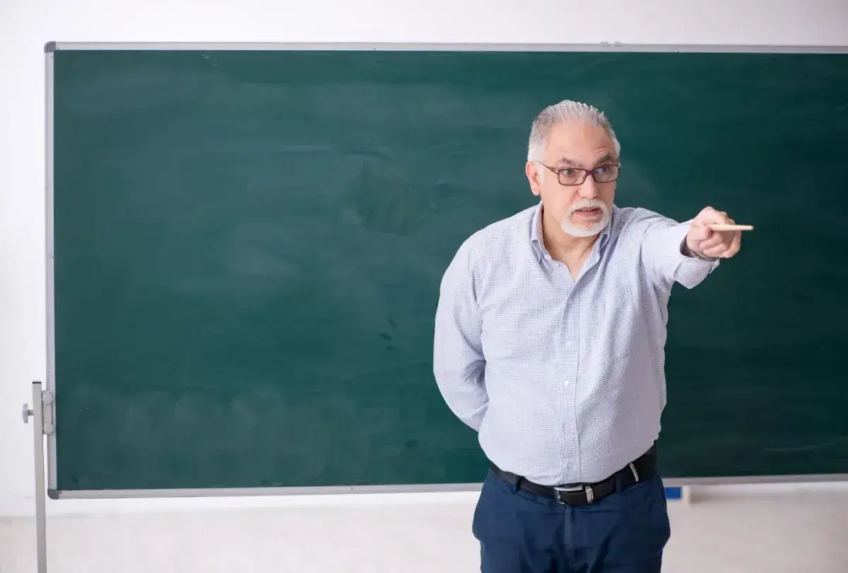 Starszy nauczyciel z kredą w dłoni, wskazujący na tablicę. Marzy o zasłużonych emeryturach dla nauczycieli.