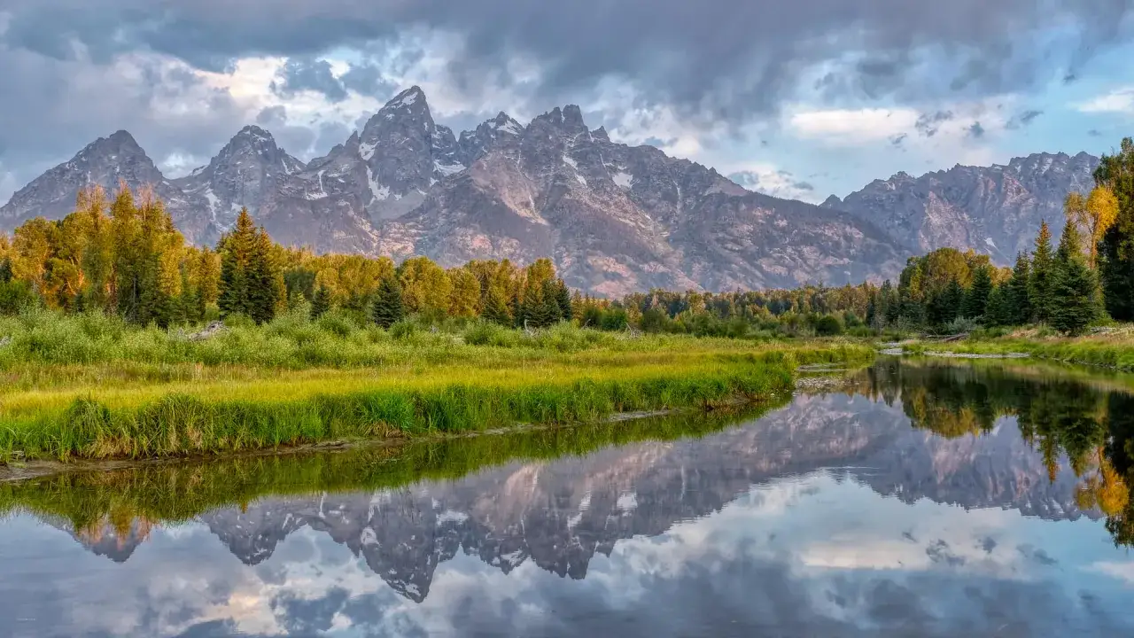 Majest&auml;tiska bergssiluetter speglas i en lugn flod, omgivna av frodig gr&ouml;nska och tr&auml;d. En vacker vy med **high dynamic range** f&aring;ngar detaljer i b&aring;de ljusa och m&ouml;rka partier.