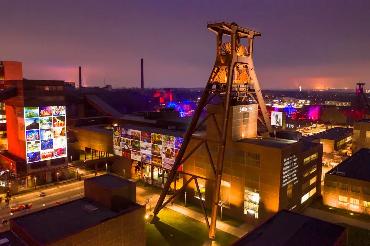 Die Zeche Zollverein nachts, beleuchtet mit bunten Projektionen auf den Gebäuden und dem Förderturm. Ein beeindruckendes Industriedenkmal im Abendlicht.