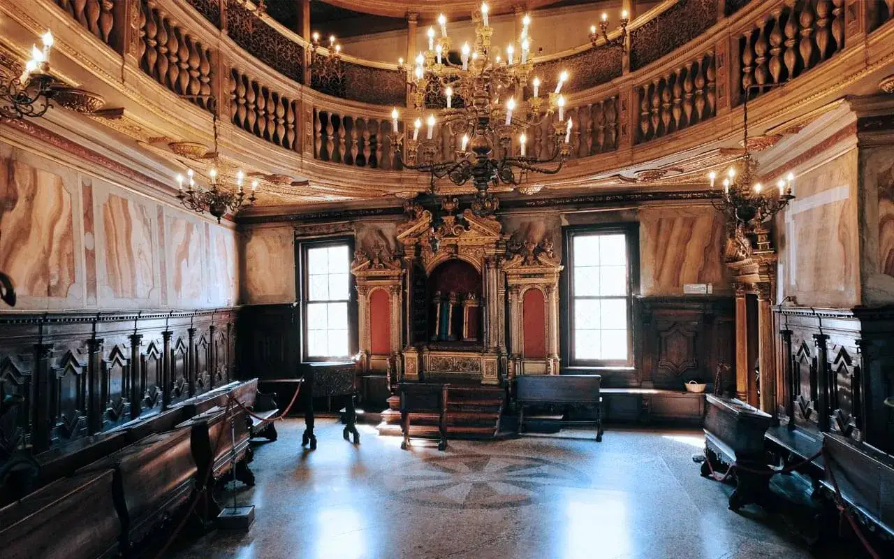 Interno sontuoso della sinagoga di Venezia, con arredi in legno scuro, un grande lampadario e l'Arca Santa dorata.