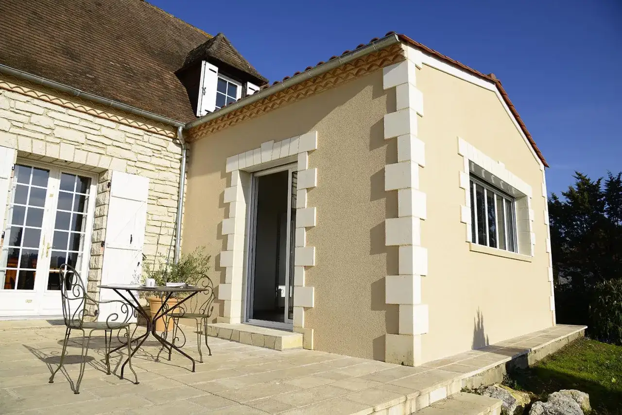 Extension de maison sur terrasse béton. Une table et des chaises invitent à profiter du soleil.