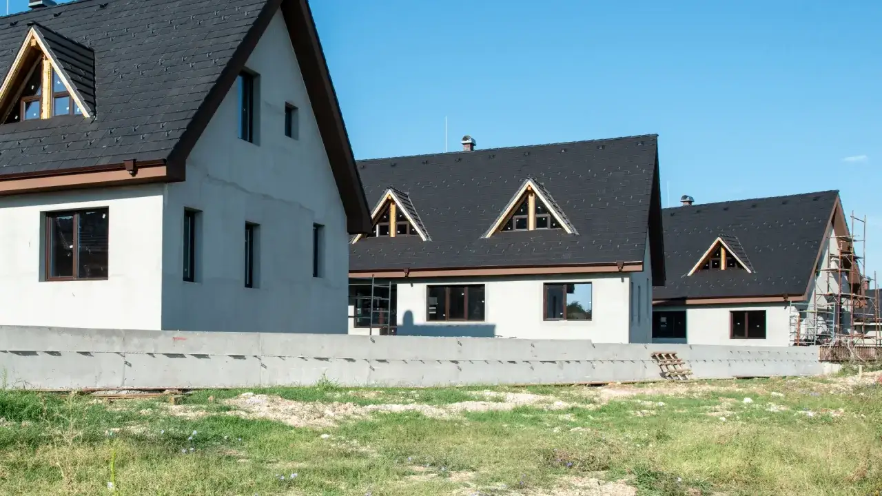 Nowe domy w budowie, z ciemnymi dachami i jasnymi ścianami, czekają na wykończenie. Dachówka skutecznie chroni ścianę przed deszczem.