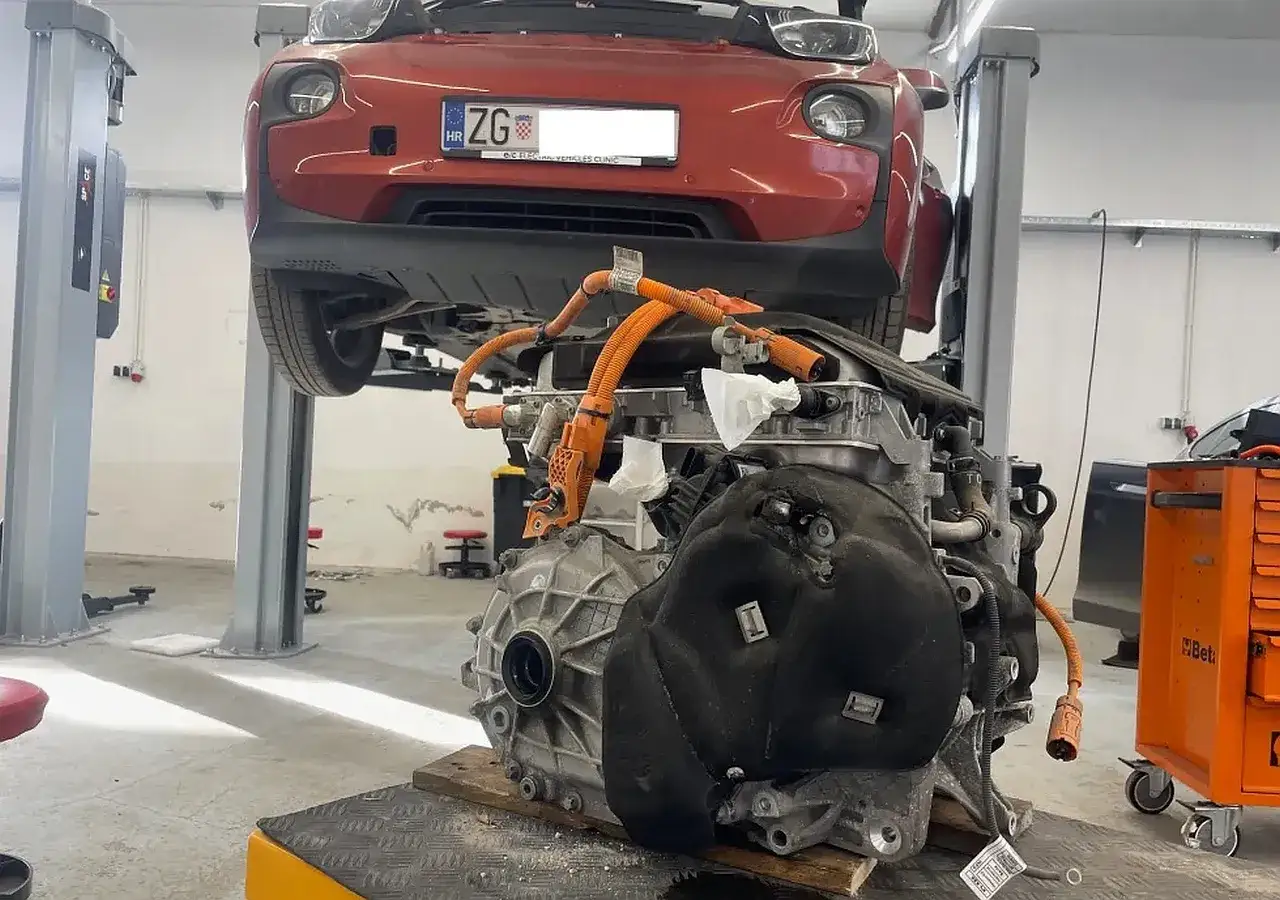 Czerwony samoch&oacute;d na podnośniku, pod nim wyjęty silnik elektryczny. Trwa **zatarty silnik naprawa**.