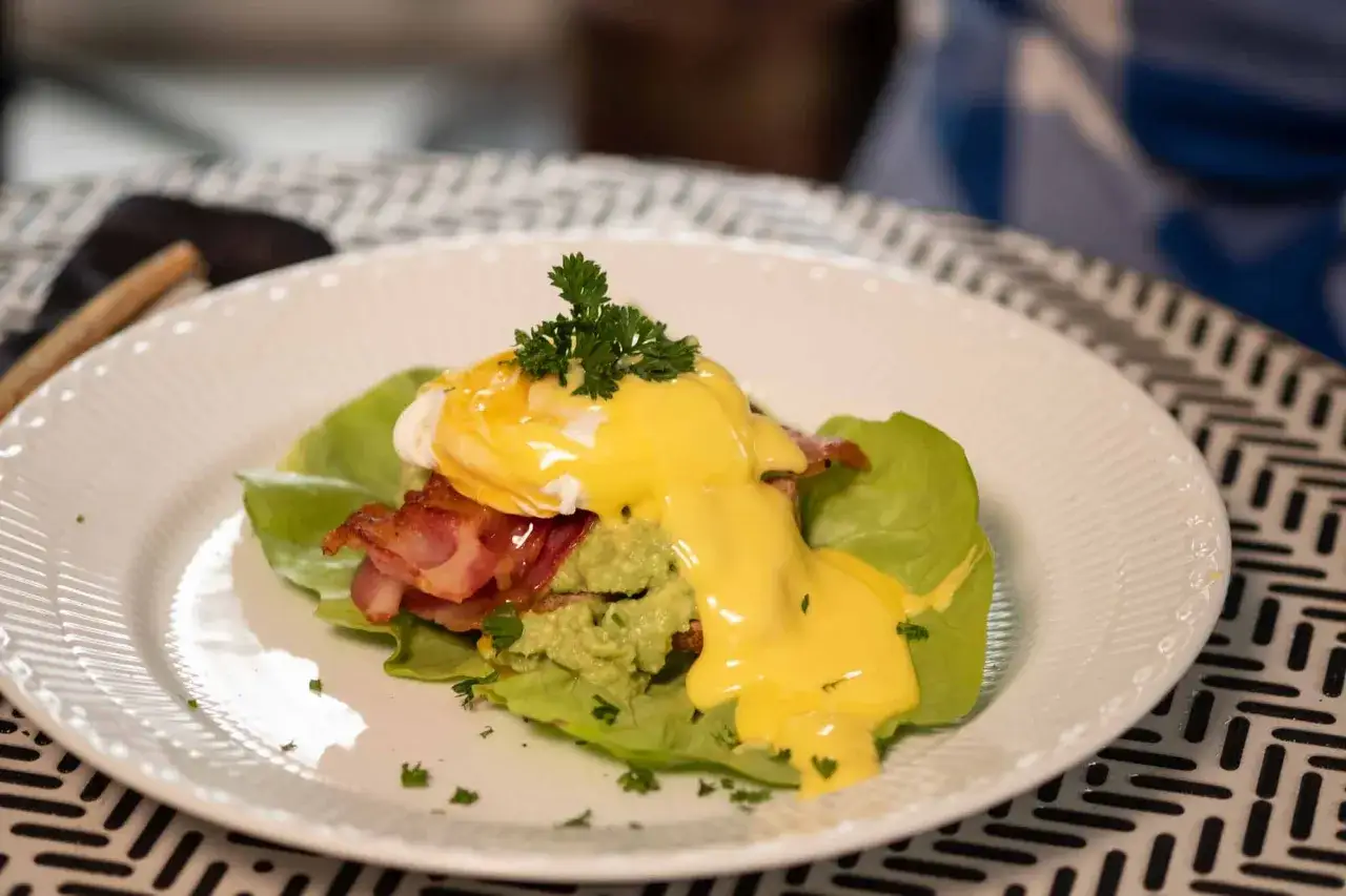 Deliciosos huevos benedictinos con aguacate y tocino crujiente, bañados en salsa holandesa y decorados con perejil fresco.