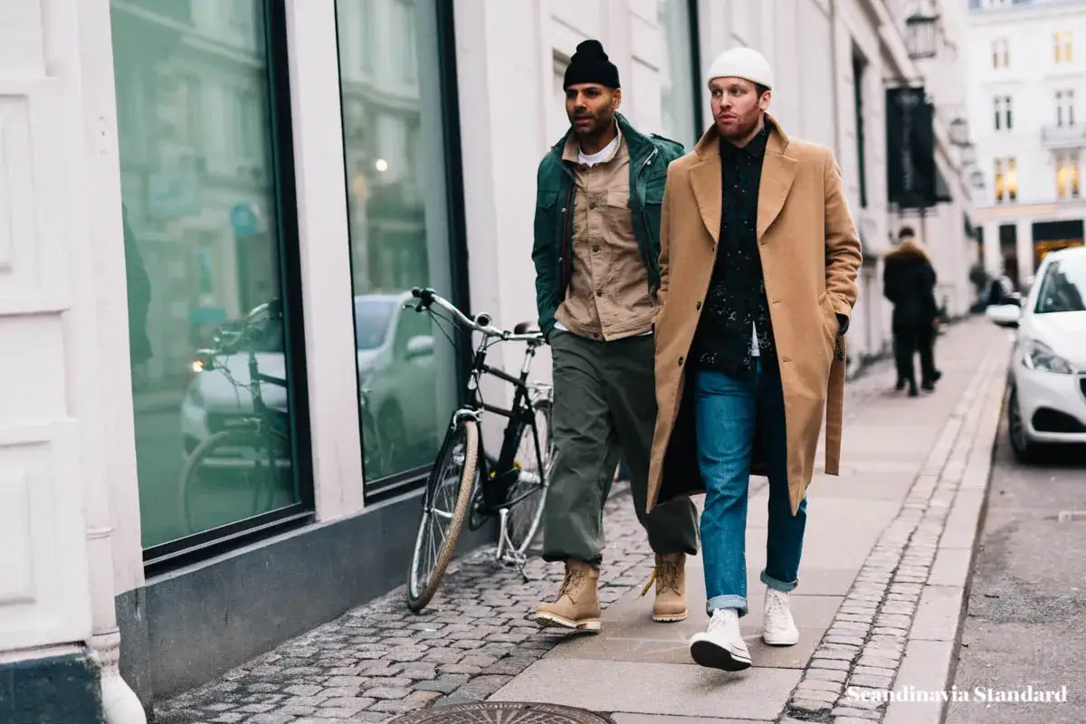 Zwei M&auml;nner spazieren mit einem Fahrrad. Sie tragen l&auml;ssige Outfits, die typisch f&uuml;r skandinavische Modelabels sind: ein Mann im beigen Mantel, der andere in einer gr&uuml;nen Jacke.