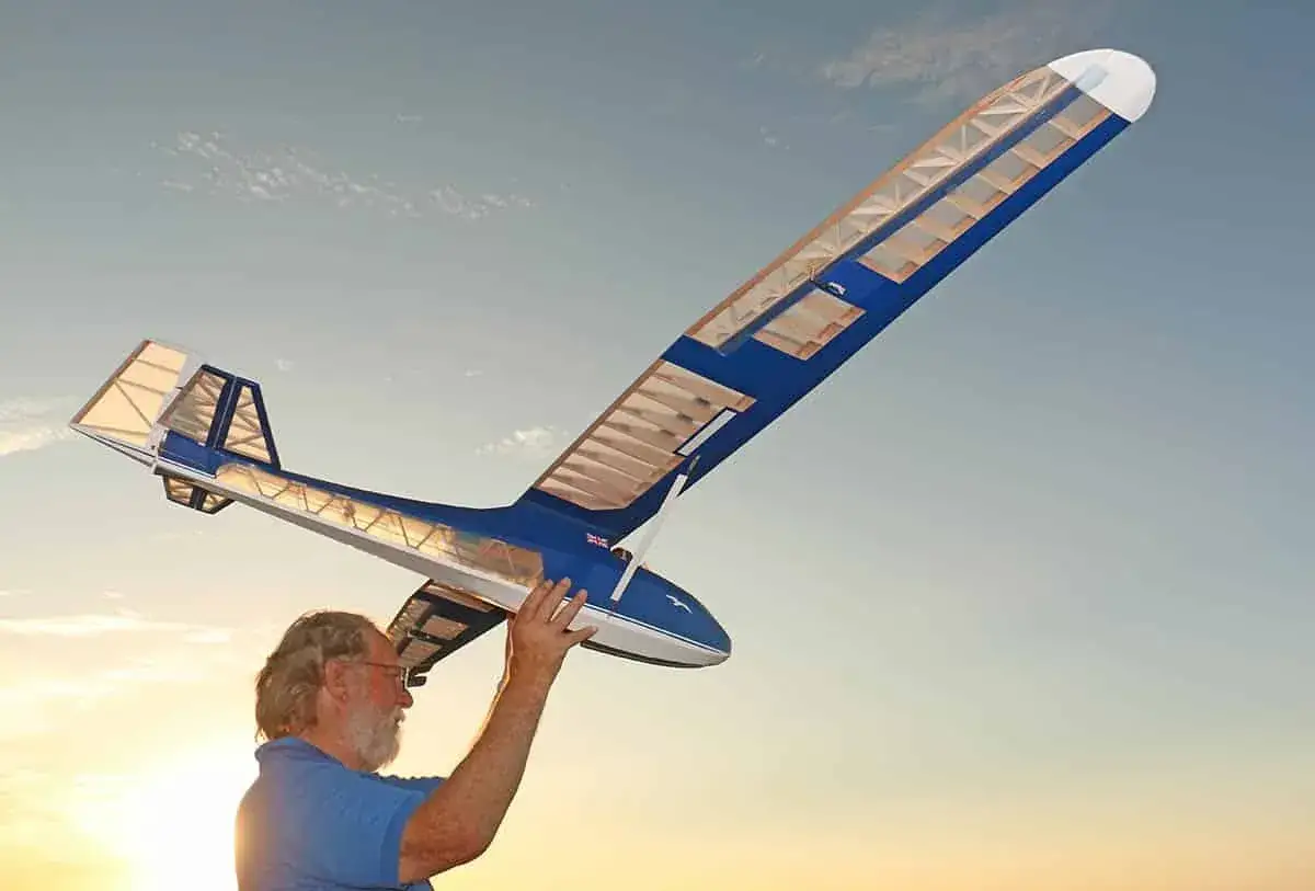 Ein Mann hält ein selbstgebautes Segelflugzeug in den Händen. Der Himmel ist blau mit wenigen Wolken.