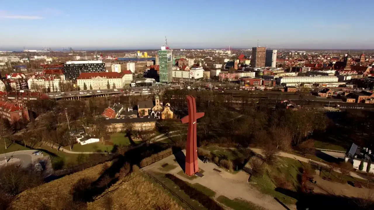 Widok na miasto z czerwonym krzyżem na pierwszym planie. W tle widać budynki, w tym wieżowce, oraz fragment portu. To teren dawnego fortu G&oacute;ry Gradowej.