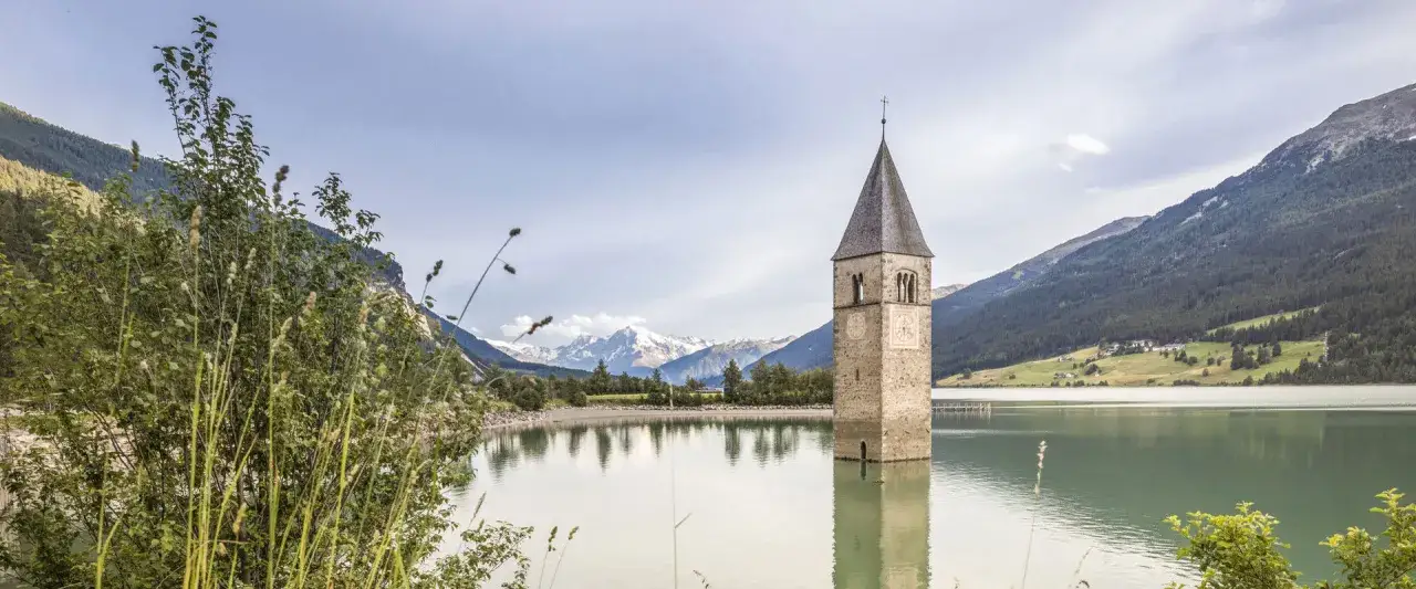 Torre di un antico campanile emerge da un lago alpino, circondato da montagne verdi e innevate.