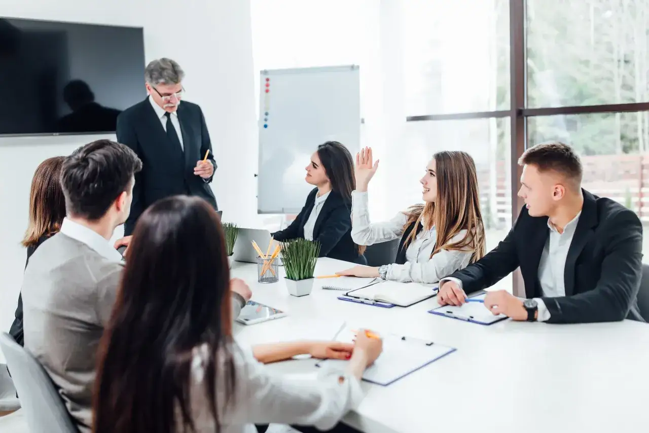 Un uomo in giacca e cravatta parla a un gruppo di giovani professionisti durante un **one to one meeting**. Una ragazza alza la mano per fare una domanda.