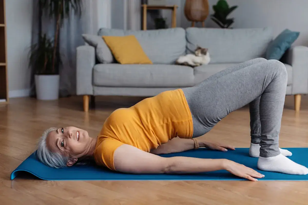 Eine Frau mit grauem Haar macht eine Brücke auf einer blauen Matte. Sie trägt ein orangefarbenes Oberteil und graue Leggings. Ihr Lächeln strahlt einen tollen Vibegron aus.