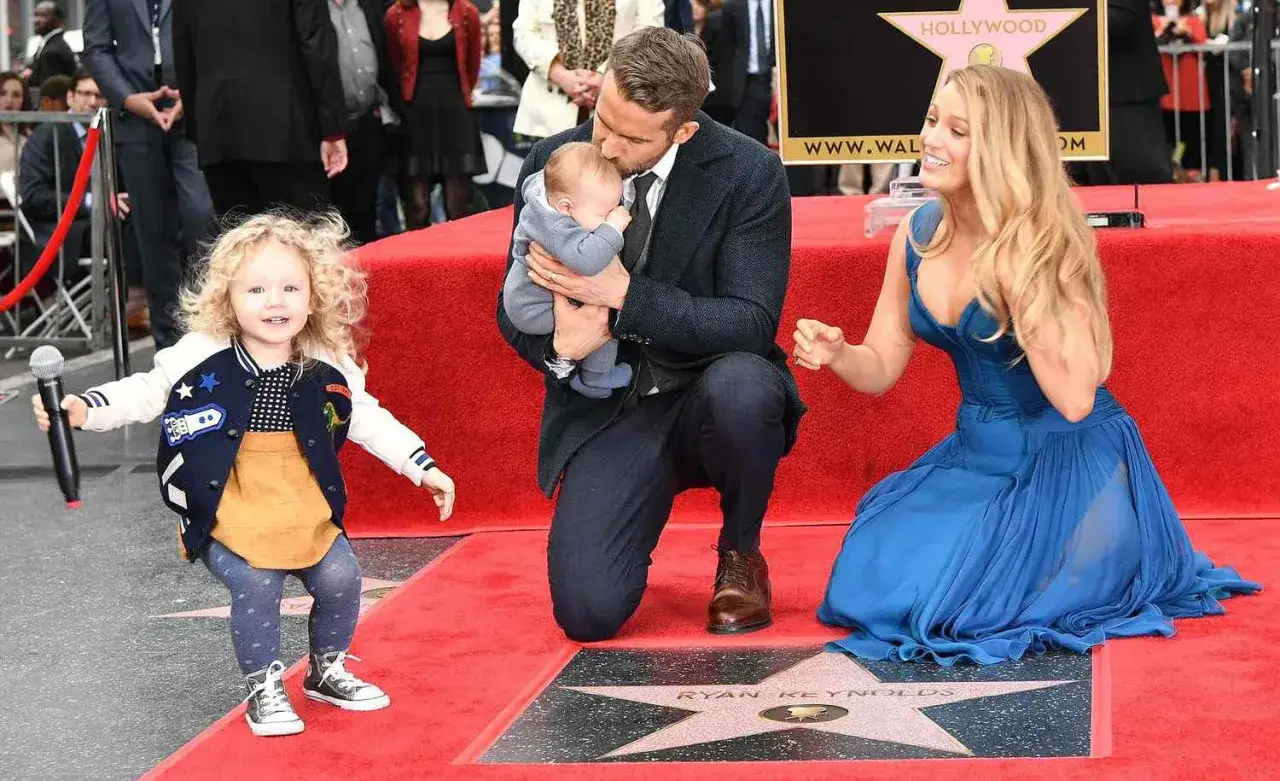 Blake Lively z dziećmi na czerwonym dywanie. Ryan Reynolds trzyma niemowlę, a starsza córka, James, bawi się mikrofonem.