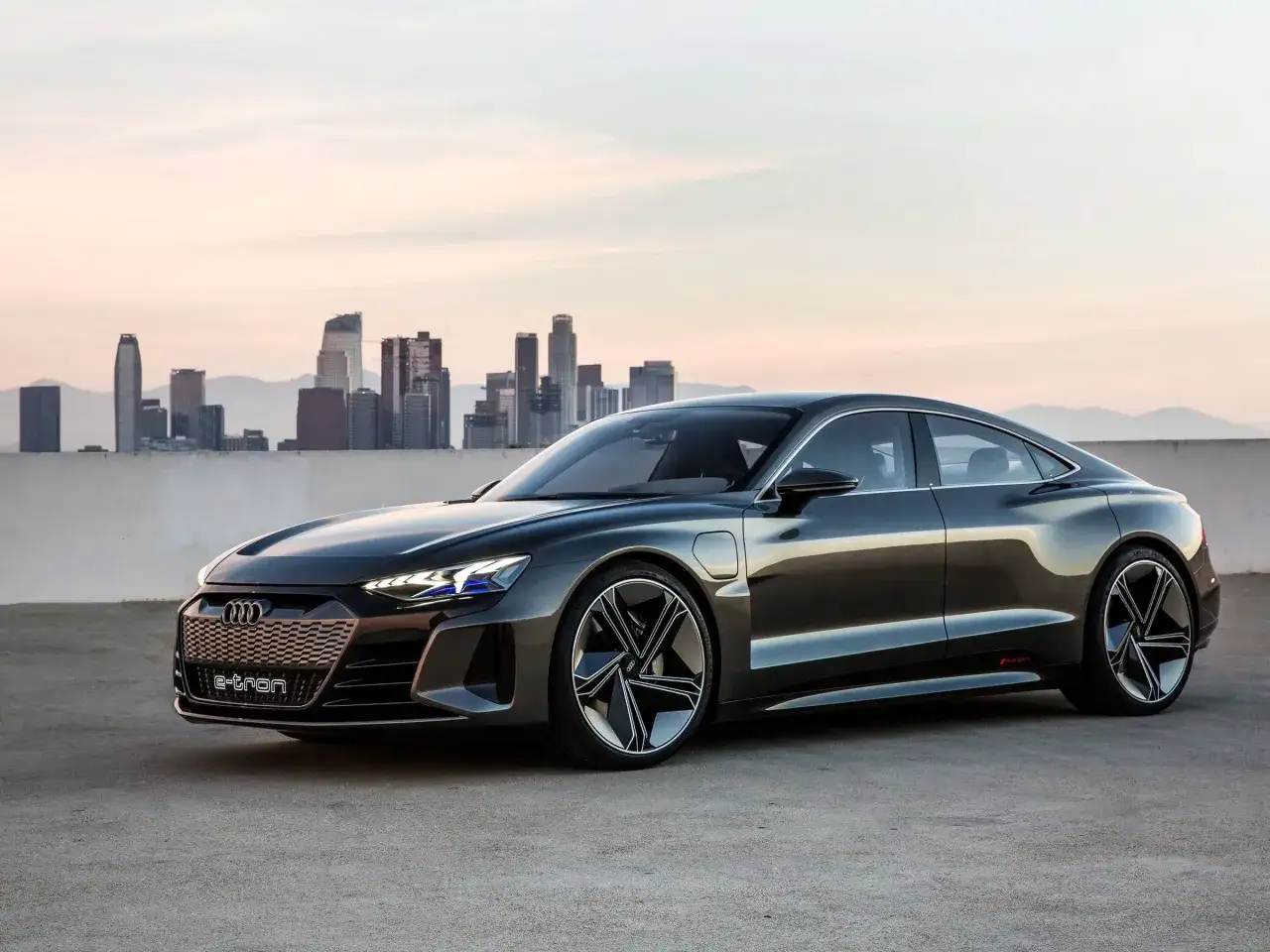 Elegancki, ciemnoszary Audi e-tron GT stoi na tle panoramy miasta o zachodzie słońca.