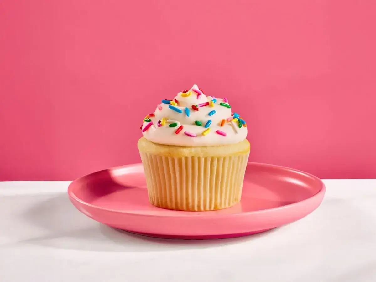 Ein Cupcake mit weißem Zuckerguss und bunten Streuseln als **cupcake topping** auf einem rosa Teller vor pinkem Hintergrund.