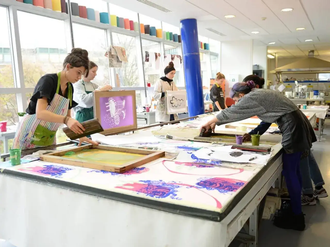 Des étudiants apprennent le métier d'artiste en sérigraphie, manipulant des cadres et des encres pour créer des motifs colorés sur du tissu.