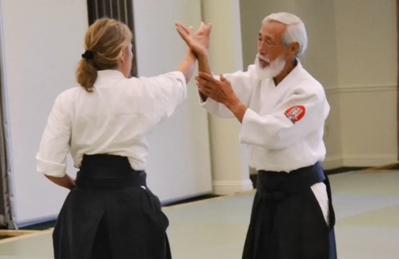 Två personer i vit gi tränar kampsport aikido. En äldre man med vitt skägg och en kvinna med hästsvans.