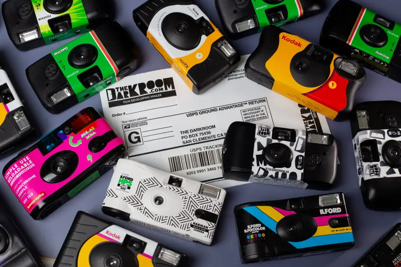 A collection of colorful disposable cameras, including Kodak, Fujifilm, and Ilford models, are laid out with a film developing mailer. This shows what to do with old cameras: send them for developing!