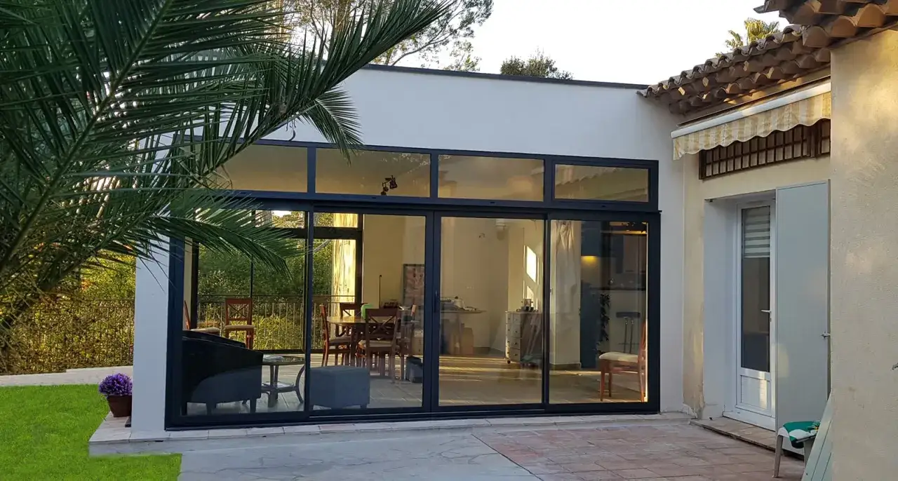 Une extension vitr&eacute;e moderne baign&eacute;e de soleil, offrant une vue sur un salon et une salle &agrave; manger. Un palmier ajoute une touche tropicale.