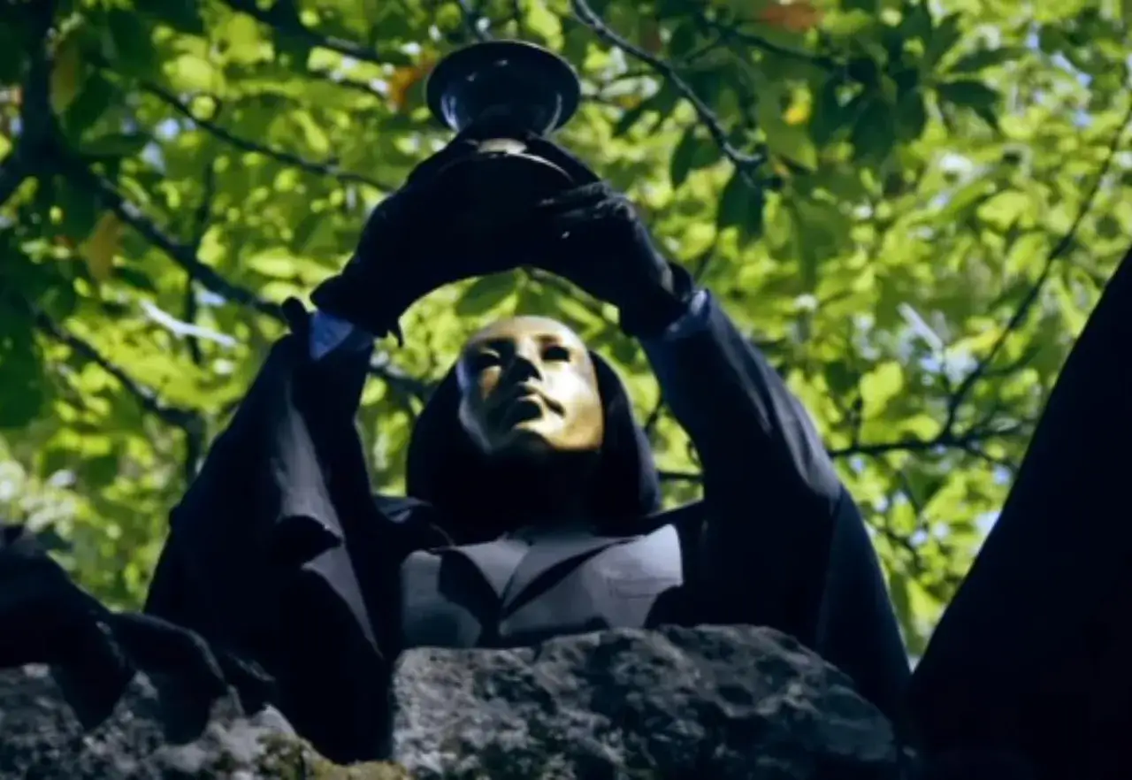 En figur med gyllene mask och svart mantel håller upp en bägare, som en hemlighetsfull scen från