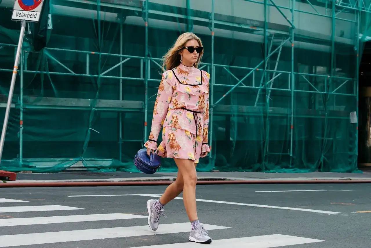 Mujer cruza la calle con vestido corto floral rosa y zapatillas deportivas grises. Un ejemplo de que zapatos usar con vestido corto.