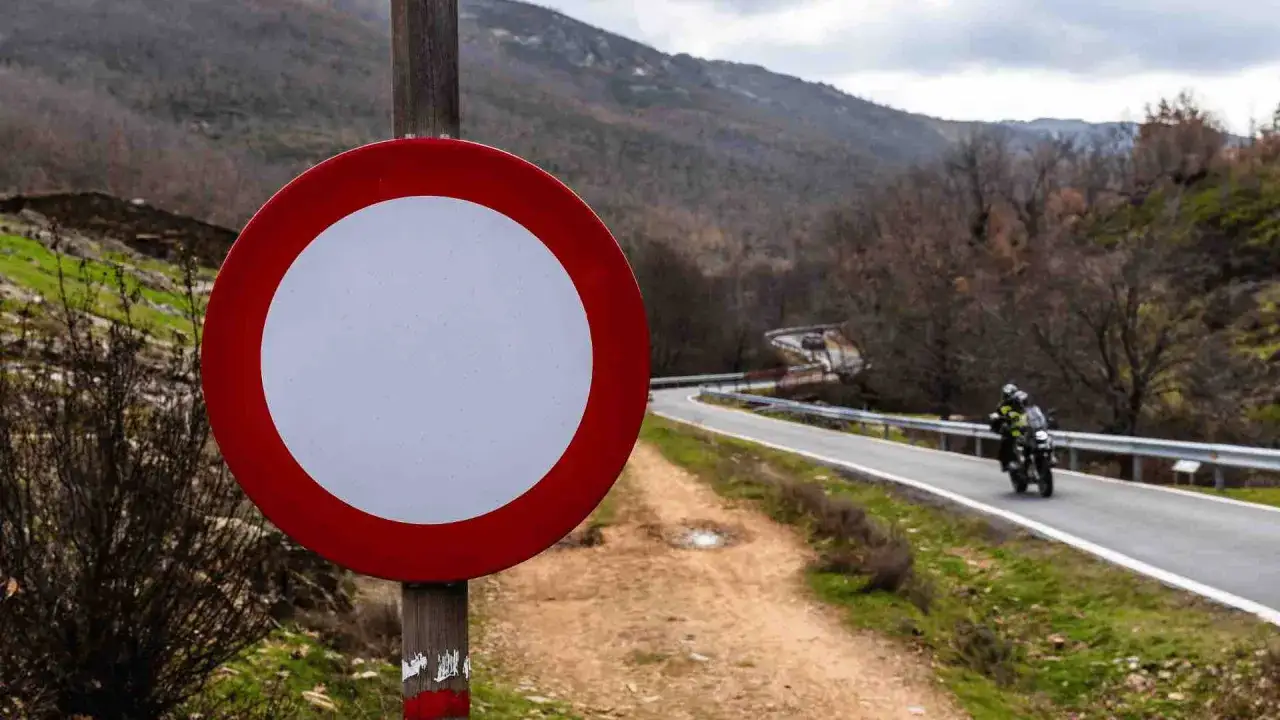 Panneau sens interdit sur un chemin de terre, avec une route sinueuse et des motards en arrière-plan.