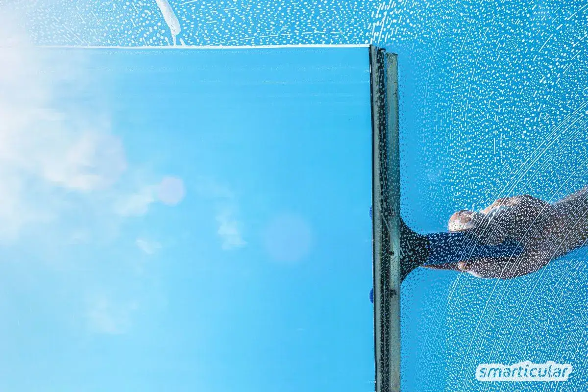 Fensterputzen mit Glycerin: Ein klarer Blick auf den blauen Himmel, w&auml;hrend eine Hand mit einem Abzieher &uuml;ber die schaumbedeckte Scheibe gleitet.
