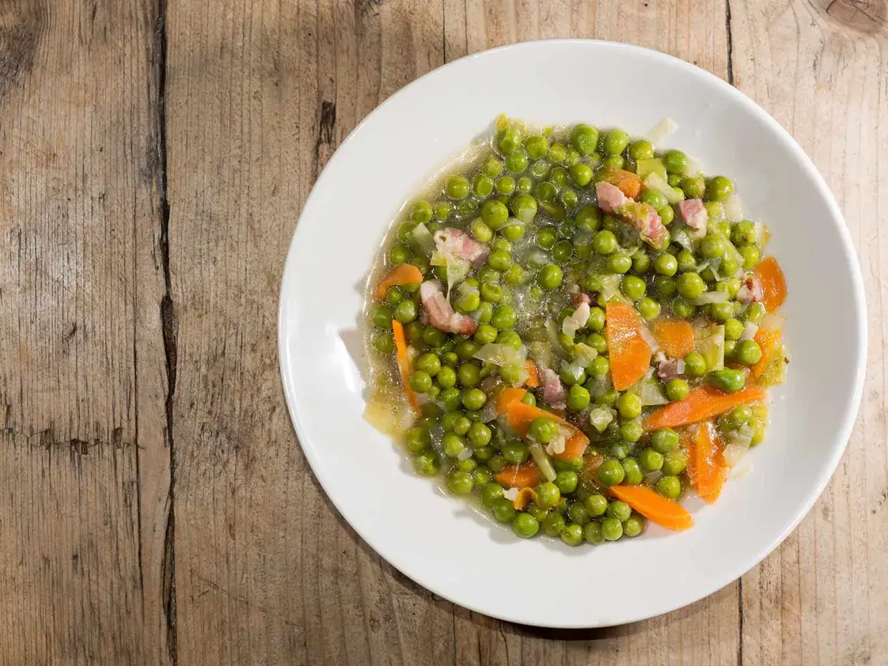 Un plat de petit pois carotte, avec des lardons, servi dans une assiette blanche sur une table en bois.