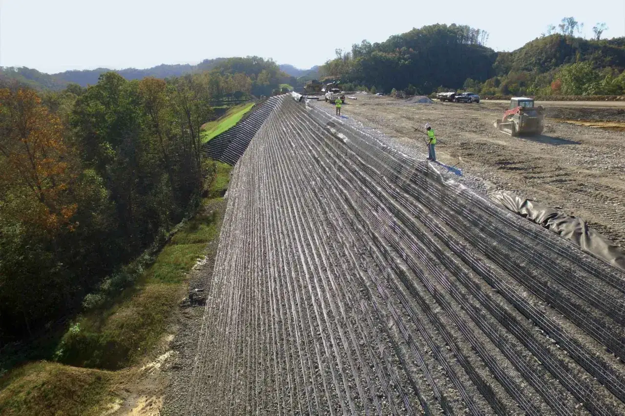 Construcci&oacute;n de talud con geomalla de refuerzo, trabajadores y maquinaria en obra.