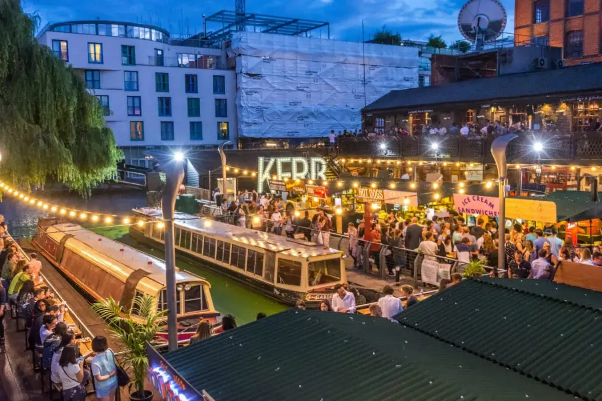 Serata vivace nei locali di Londra, con gente che passeggia lungo il canale e si gode l'atmosfera festosa tra bancarelle e bar illuminati.