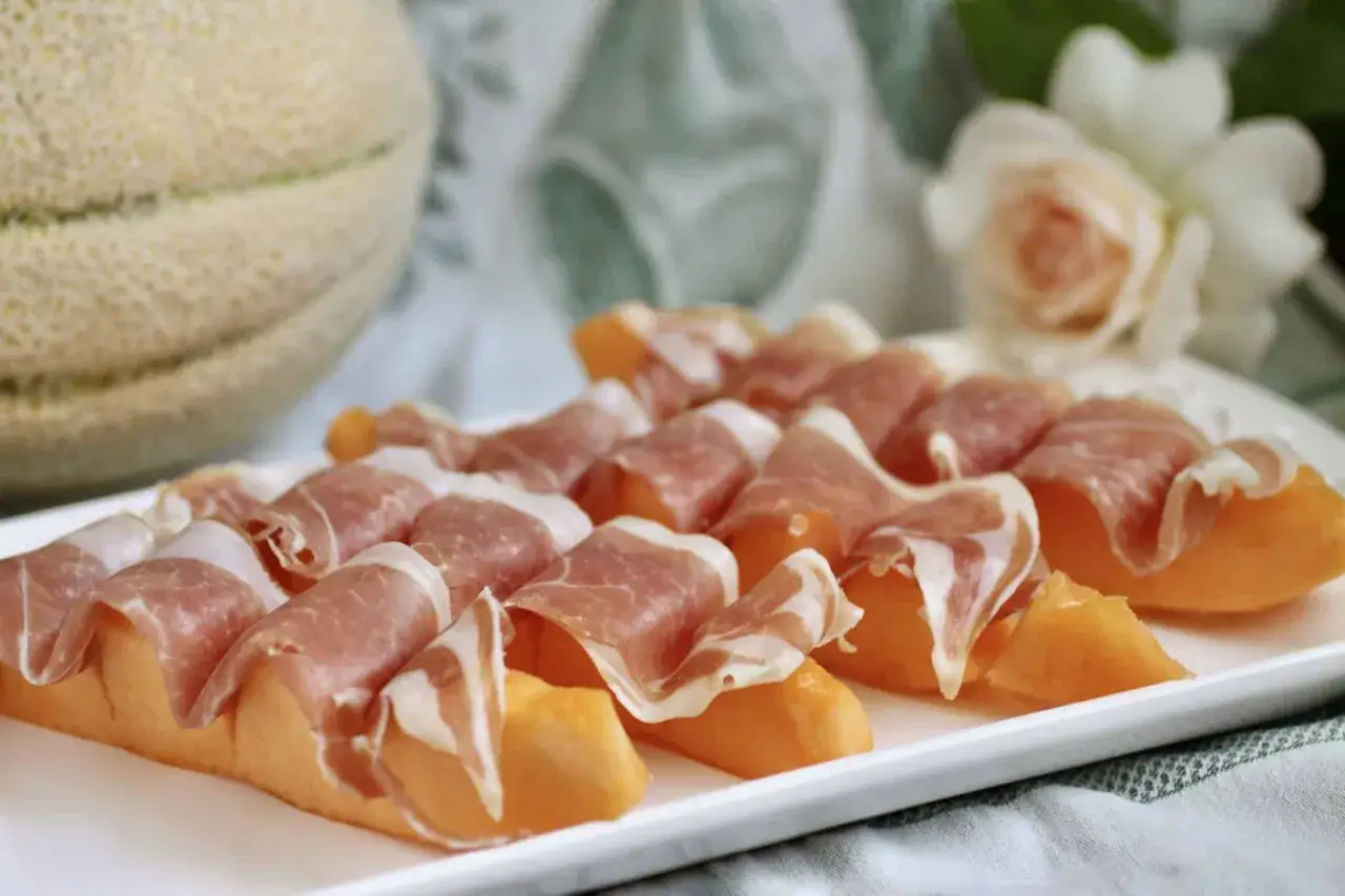 Melonbitar inslagna i tunt skivad prosciutto p&aring; en vit tallrik. En hel melon syns i bakgrunden.