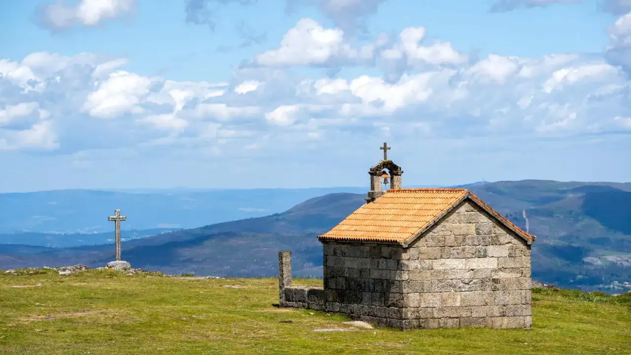 Pequeña ermita de piedra con tejado naranja y campanario en la Serra do Cando. Cruz de piedra a la izquierda.