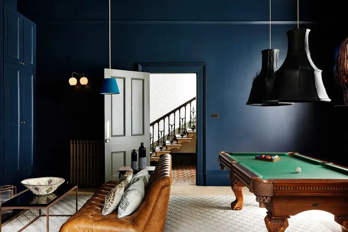 Salon avec billard et canapé en cuir marron, murs d'un profond couleur bleu foncé. Une porte ouverte révèle un escalier.