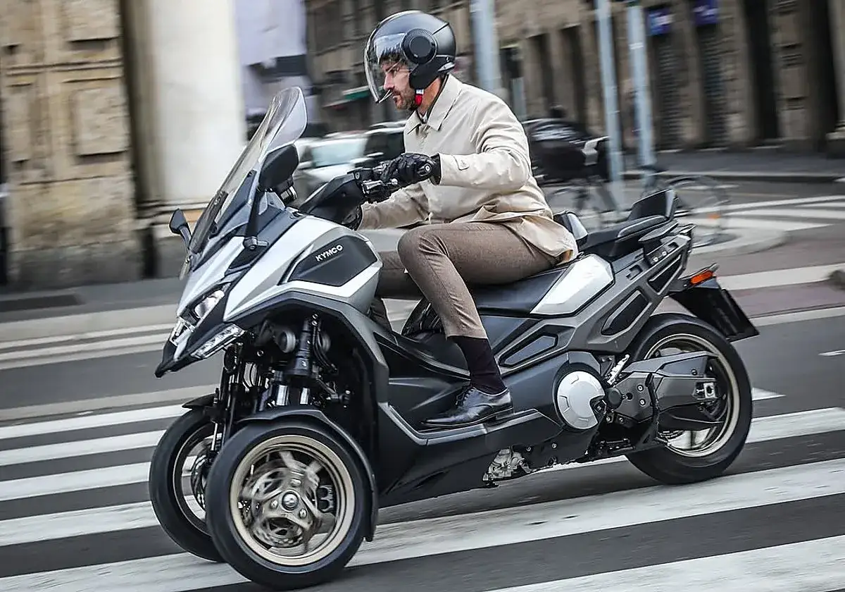 Hombre con casco conduce un triciclo Kymco, ideal para **motos para ciudad**, cruzando un paso de peatones.
