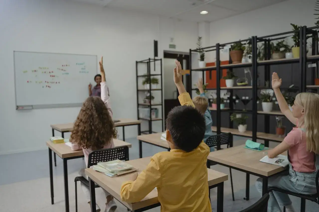 Ni&ntilde;os en un aula levantan la mano, mostrando su entusiasmo por aprender. La maestra en el frente fomenta la competencia CPSAA.