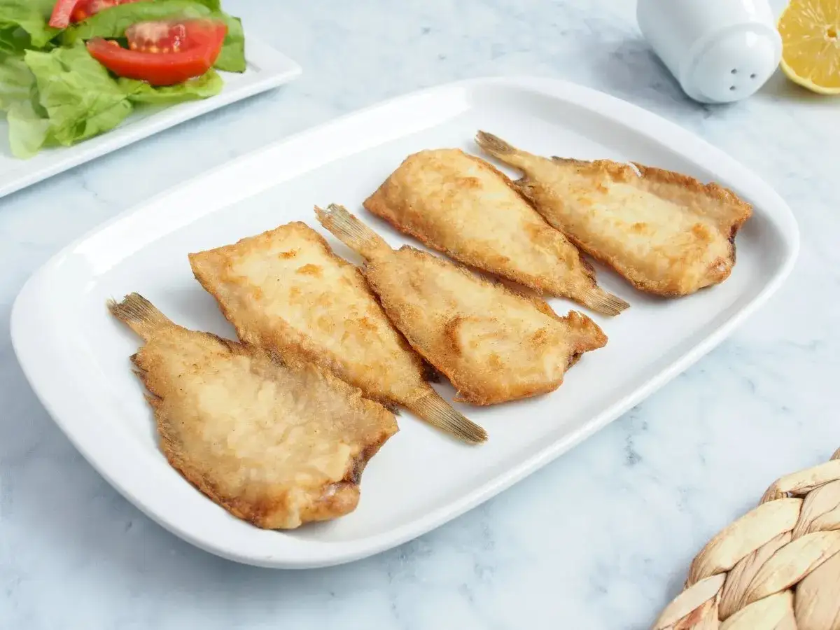 Cinco acedias fritas doradas en una fuente blanca, listas para disfrutar con una ensalada fresca.