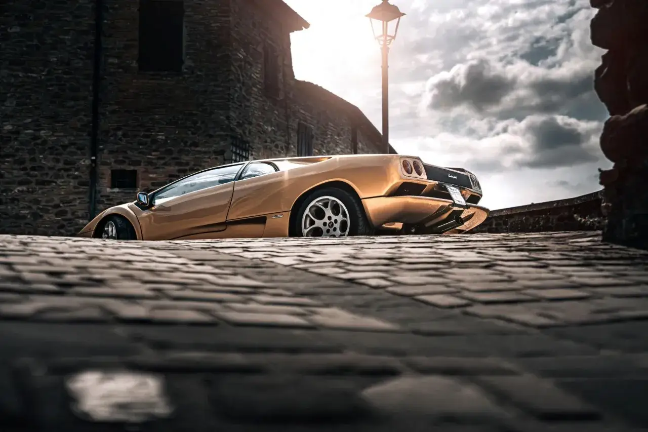 Złoty Lamborghini Diablo na brukowanej uliczce, z kamienną ścianą i latarnią w tle.