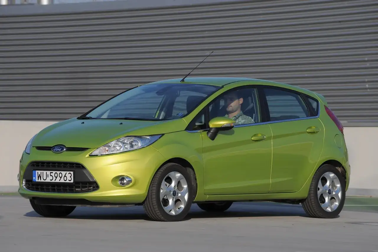 Zielony Ford Fiesta Mk7 z widocznymi żarówkami z przodu, zaparkowany przed szarą, falistą ścianą.