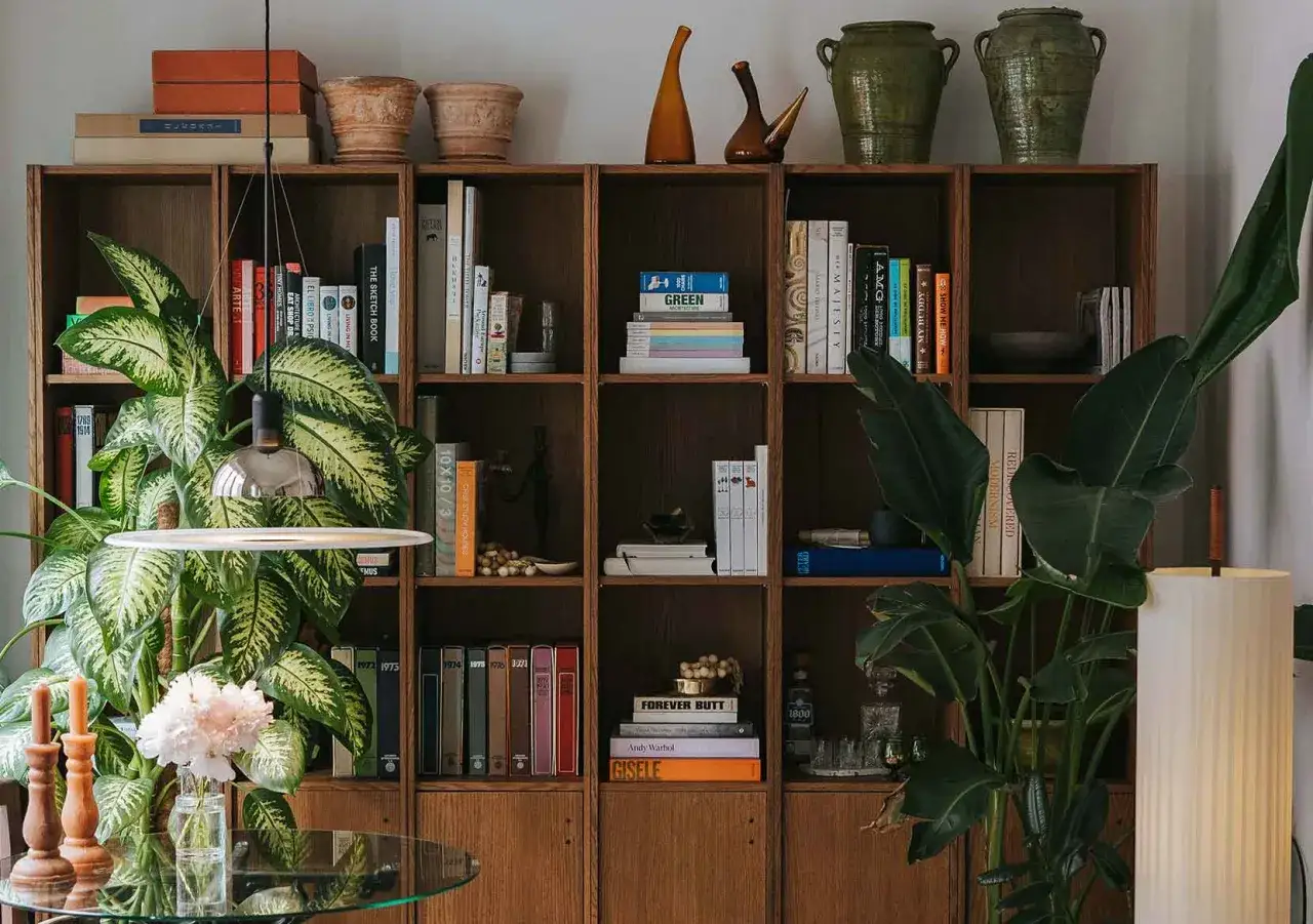 Estantería de madera con libros, plantas y objetos decorativos, creando un ambiente acogedor de despacho en casa.