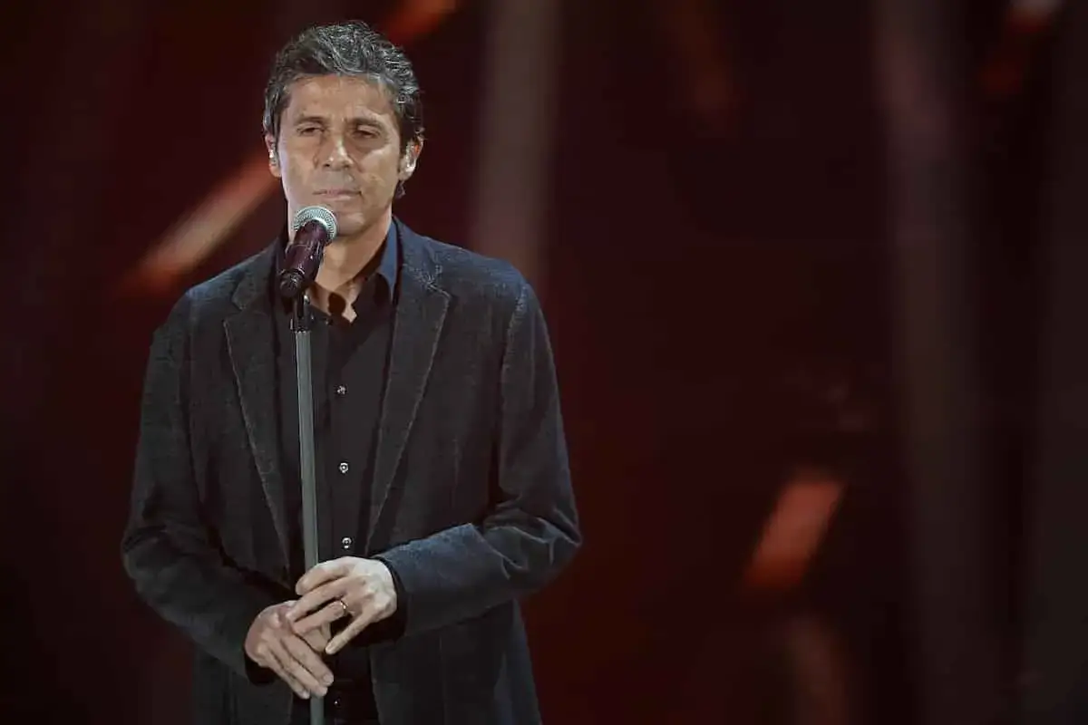 Uomo con giacca scura canta al microfono, con le mani giunte. Sembra dire