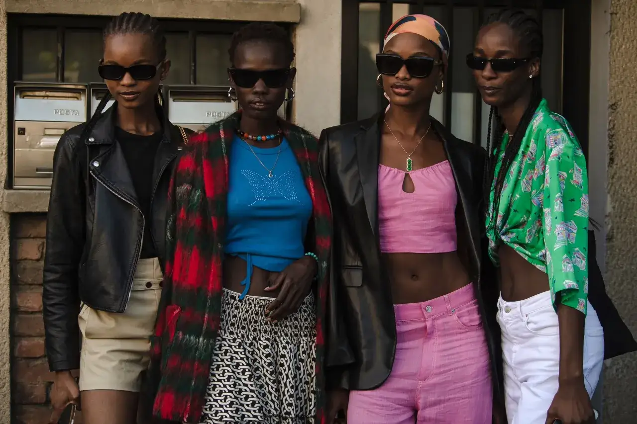 Cztery modelki w okularach przeciwsłonecznych prezentują stylizacje w klimacie y2k: skórzana kurtka, kolorowe topy, wzorzyste spódnice i spodnie.