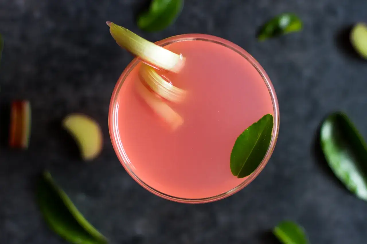 En rosa rabarbersirap drink med rabarberstavar och gr&ouml;na blad p&aring; en m&ouml;rk bakgrund.