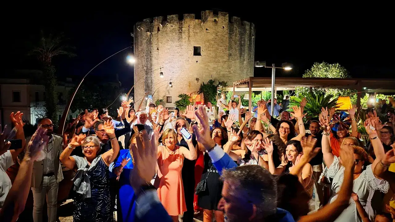 Folla festante con le mani alzate davanti a un'antica torre, un'atmosfera vibrante di eventi nel Salento.
