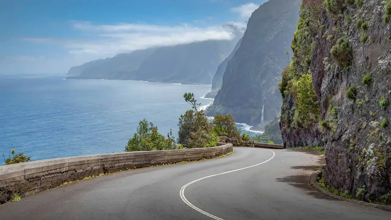 Kurvige Küstenstraße in Portugal mit Blick auf das Meer und steile Klippen. Ein Wasserfall stürzt ins Meer.