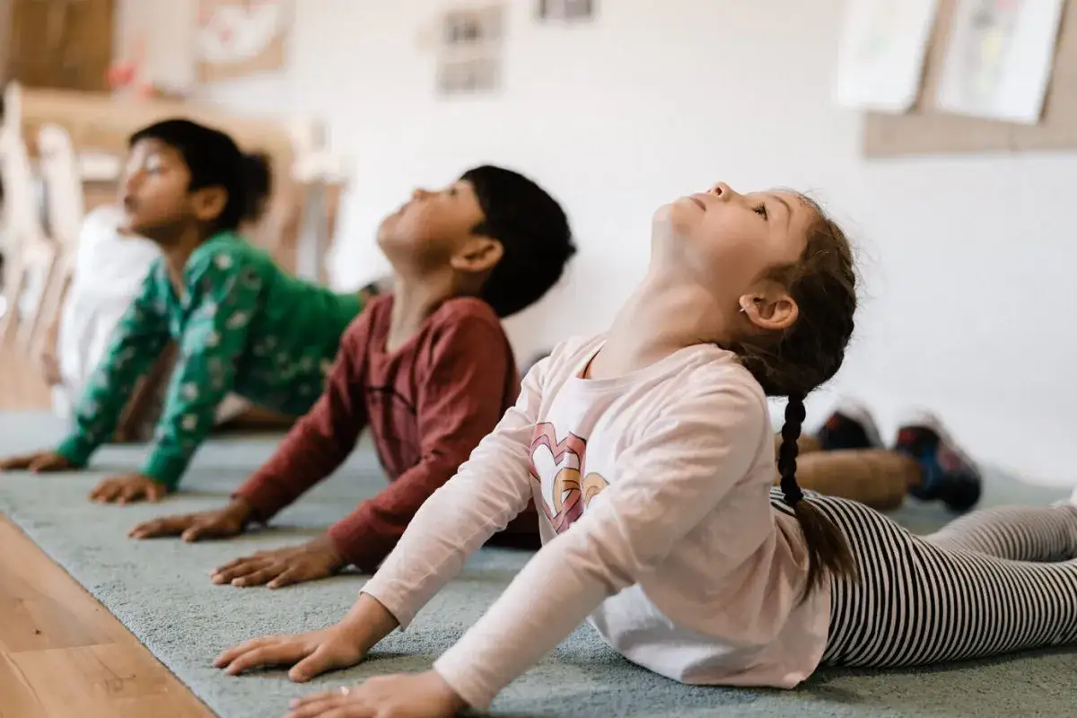 Barn gör yogaövningar på en matta. En flicka med flätat hår sträcker sig uppåt. En rolig yoga barn-stund.