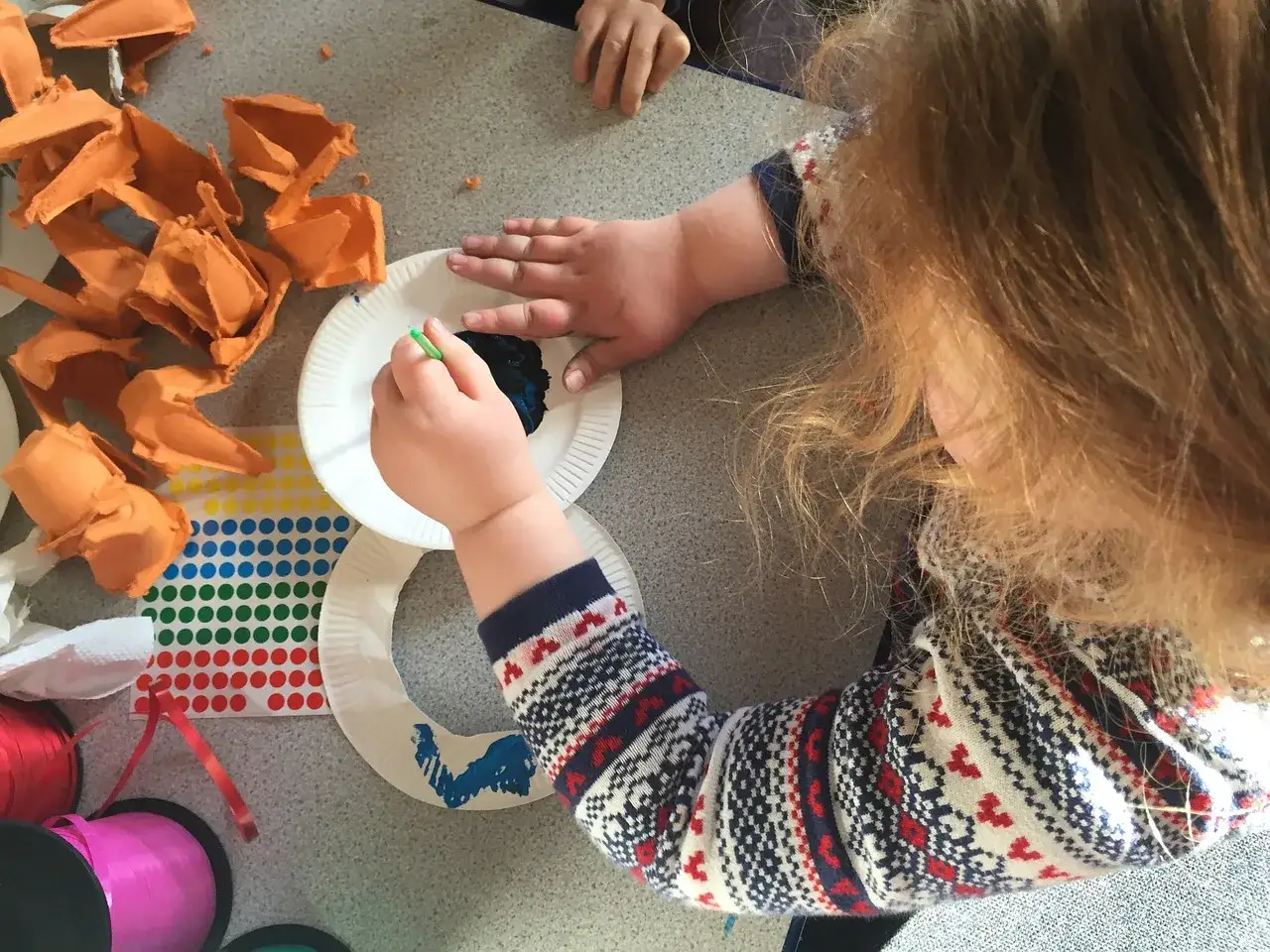 Bambini impegnati in lavori creativi: mani piccole dipingono con pennello verde su piatto bianco, circondati da materiali colorati.